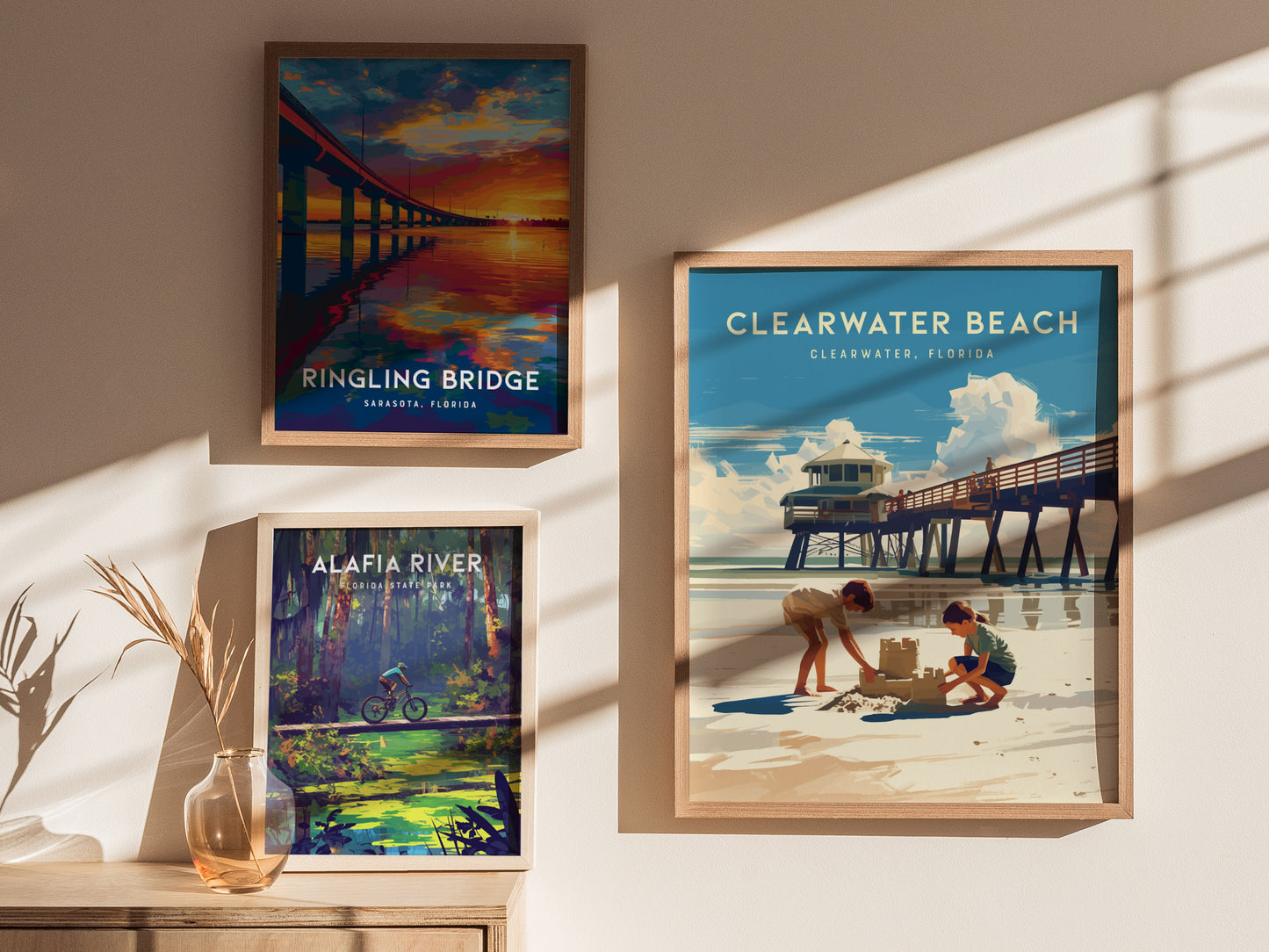 Framed posters on a wall display scenes of Ringling Bridge at sunset, Clearwater Beach with children building a sandcastle, and Alafia River with a cyclist.