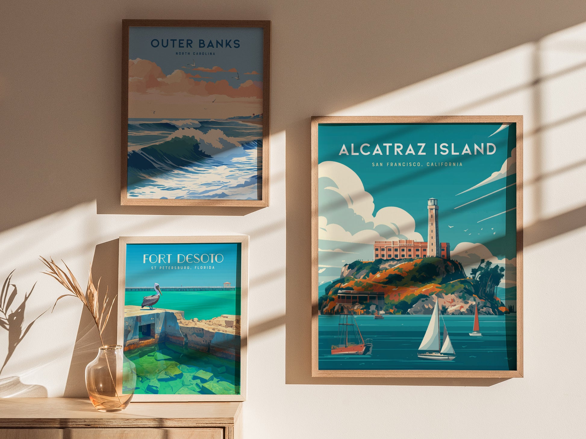 Framed wall art featuring Alcatraz Island, Outer Banks, and Fort Desoto posters displayed on a sunlit wall with a vase holding dried plants.