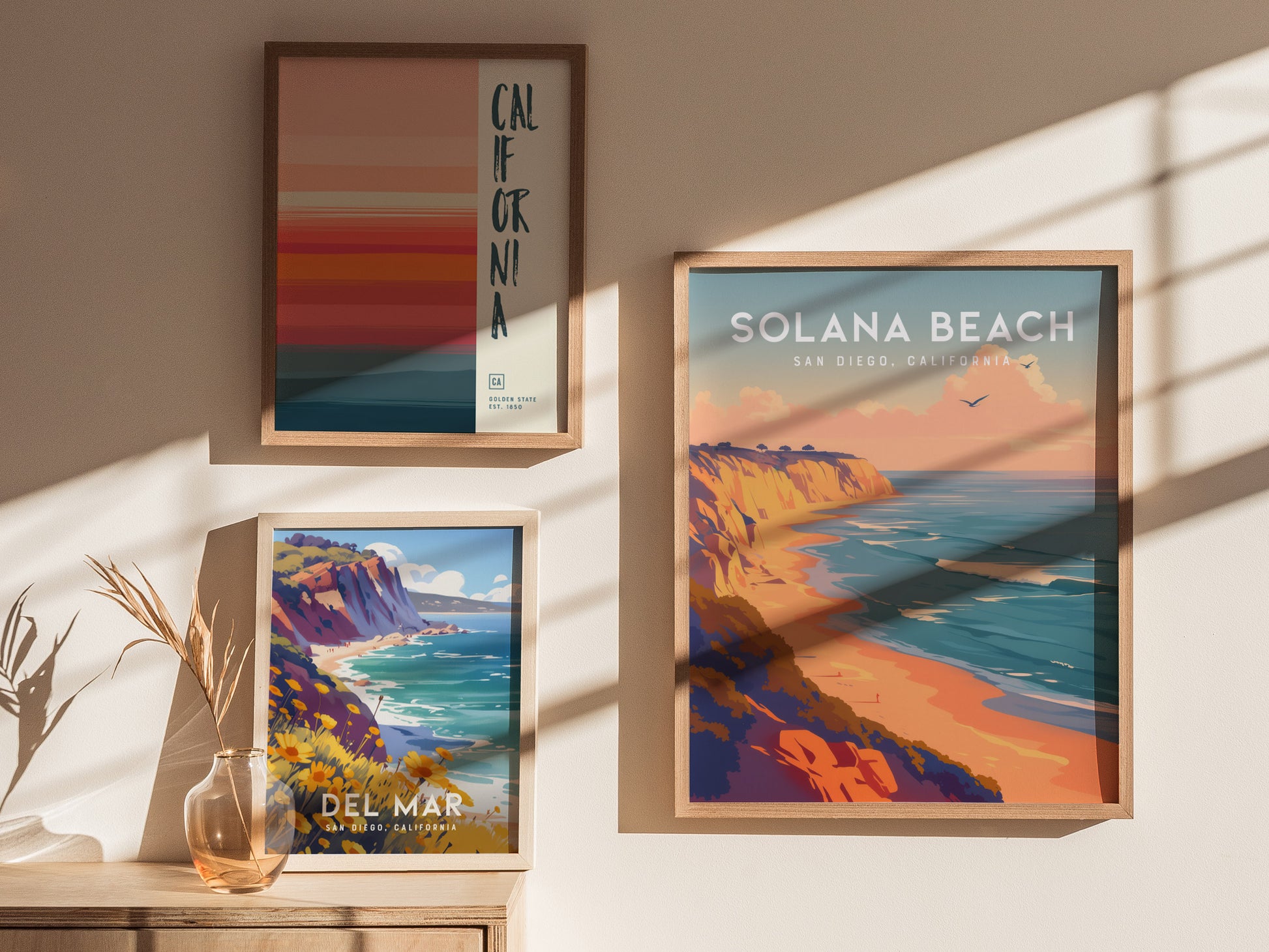 Three framed posters displayed on a wall feature vibrant coastal scenes, including Solana Beach and Del Mar, with warm tones and ocean views. Shadows and a decorative vase add depth.