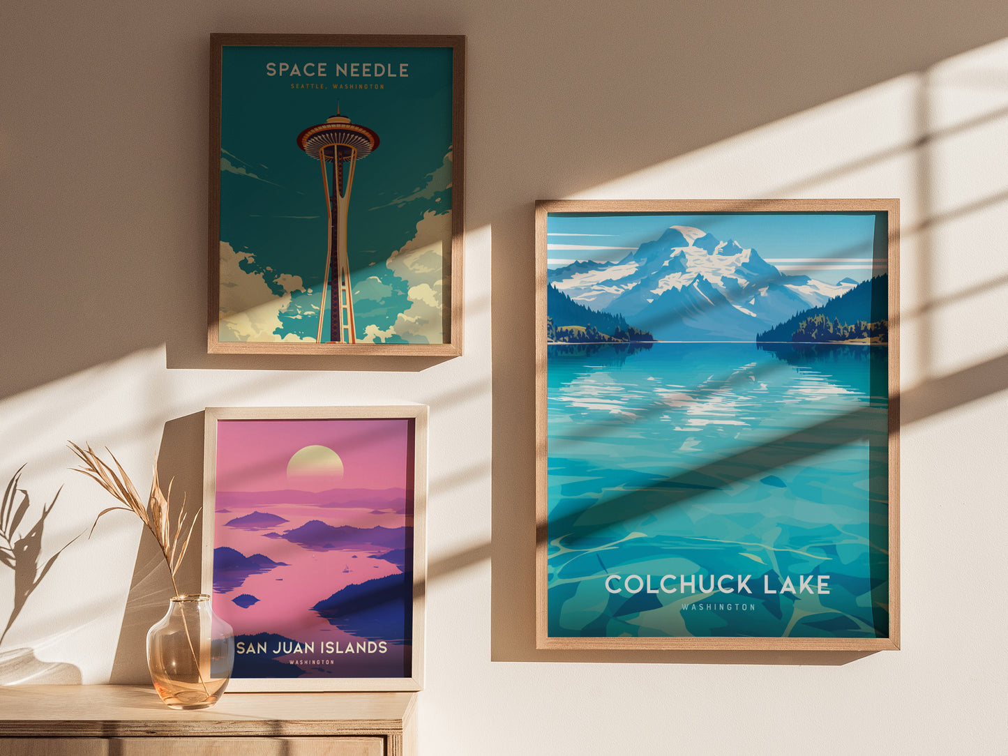 Three framed posters are displayed on a sunlit wall. They depict the Space Needle, Colchuck Lake, and the San Juan Islands, each with vibrant colors and scenic landscapes.