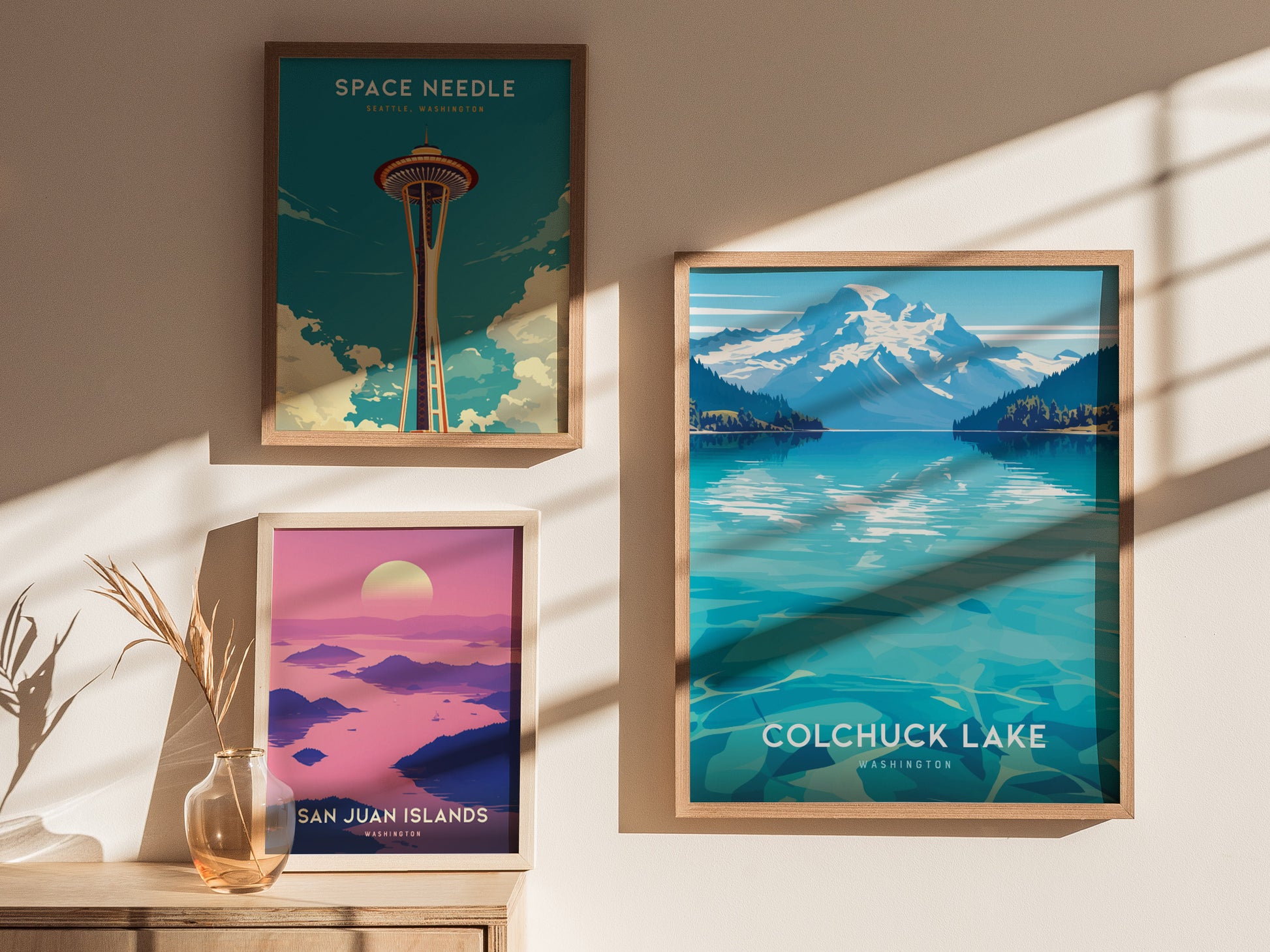 Three framed posters are displayed on a sunlit wall. They depict the Space Needle, Colchuck Lake, and the San Juan Islands, each with vibrant colors and scenic landscapes.
