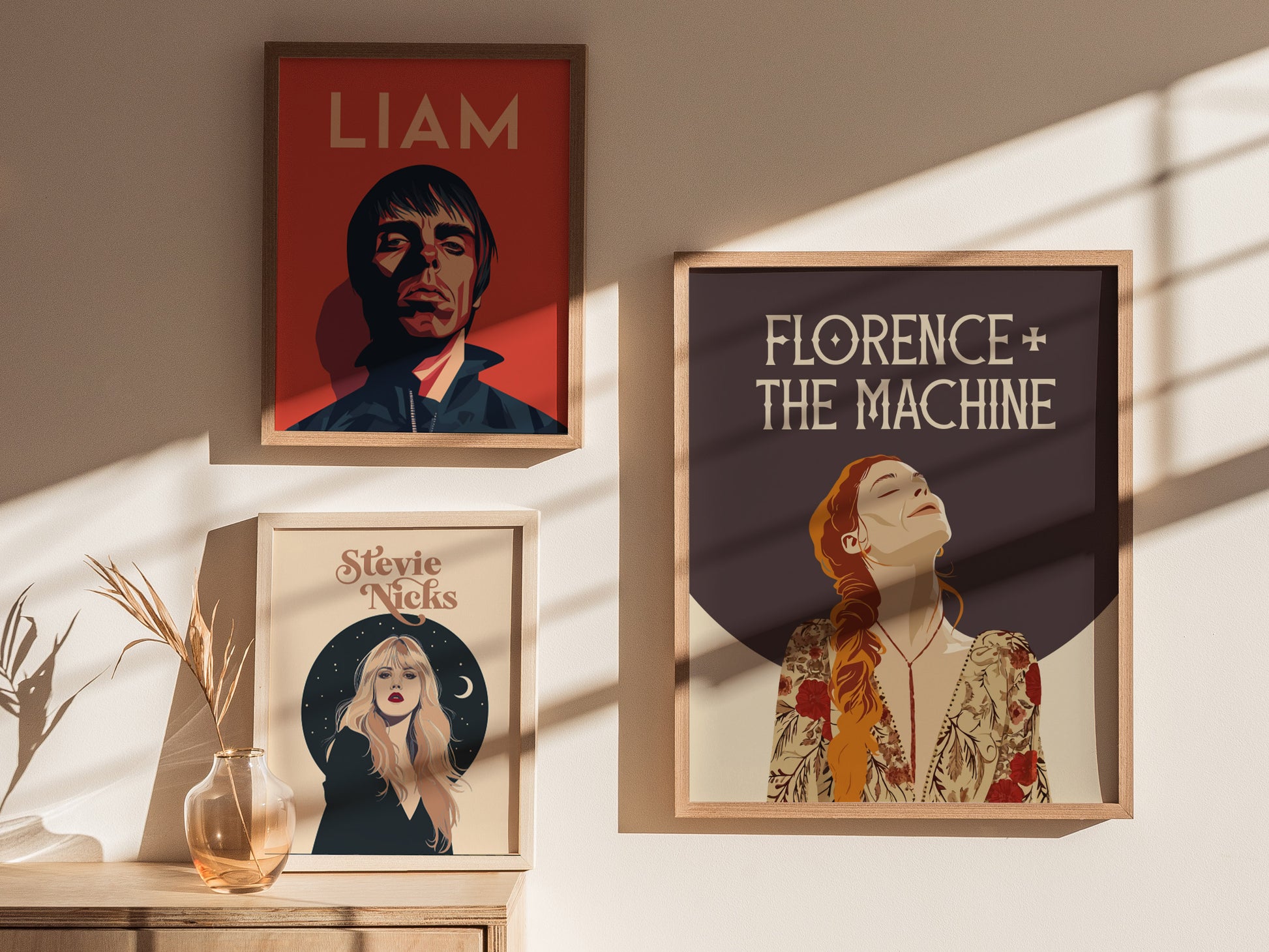 Three framed posters on a sunlit wall depict Liam Gallagher, Florence + The Machine, and Stevie Nicks, with a vase and dried plant on a wooden surface below.