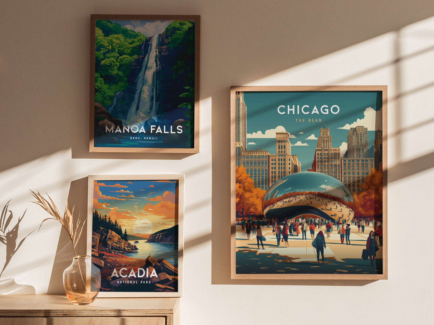 Three framed posters hang on a wall: Manoa Falls, Acadia National Park, and Chicago The Bean, with sunlight casting shadows. A vase with dried plants is on a wooden table.