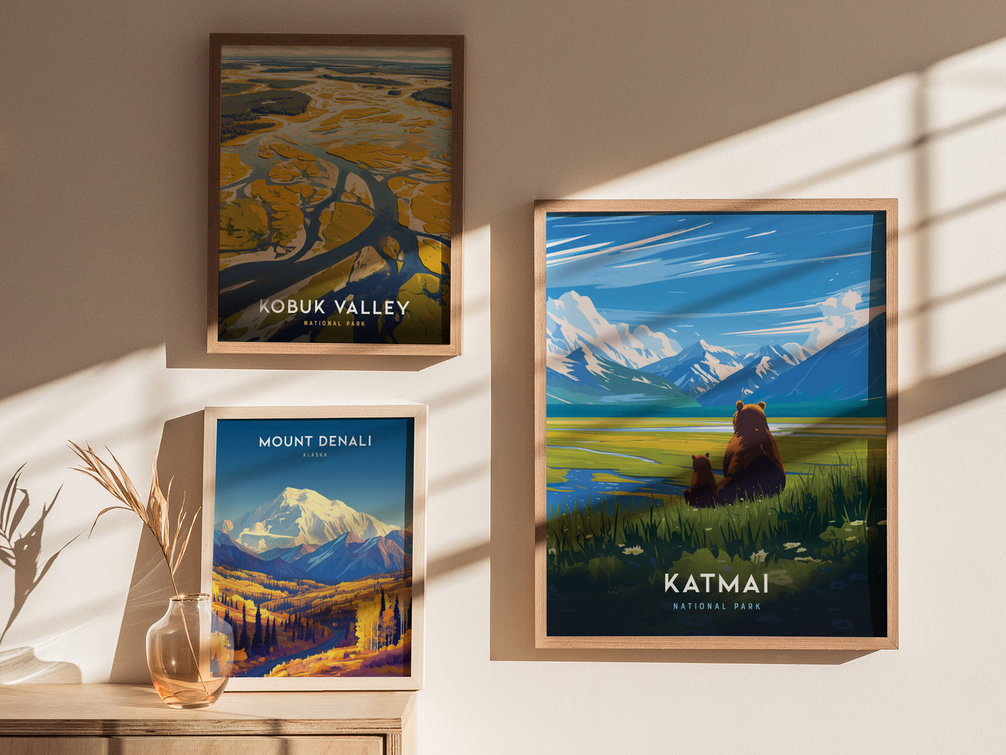 Three framed posters on a beige wall depict Kobuk Valley, Mount Denali, and Katmai National Park, featuring landscapes and wildlife, with sunlight casting shadows across them.