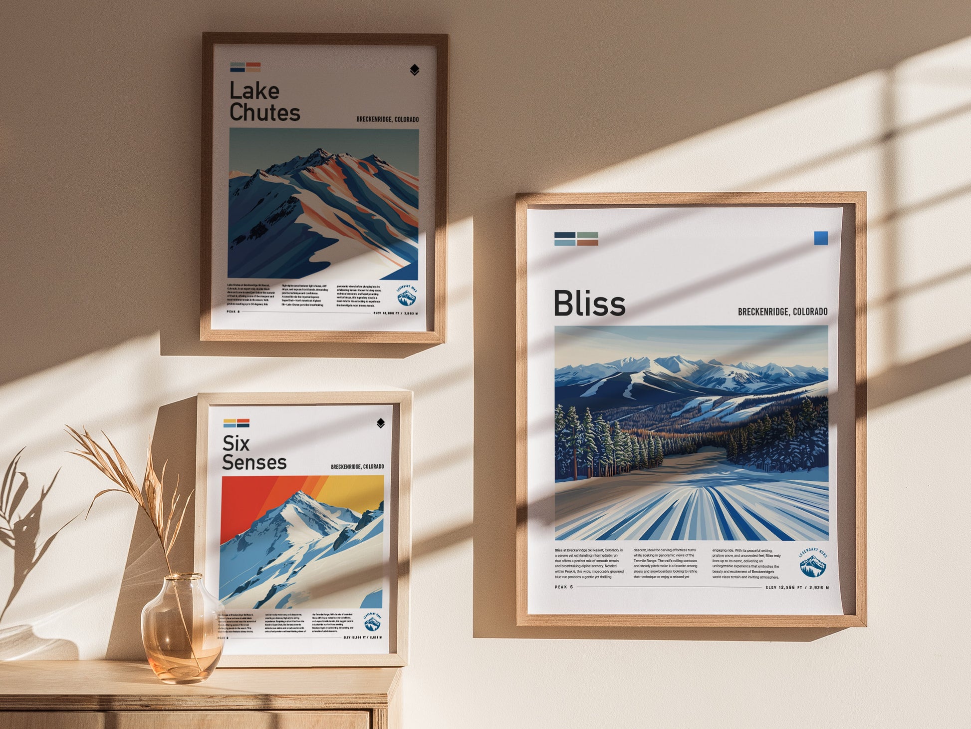 Framed wall art posters depict ski runs at Breckenridge, Colorado, with stylized mountain landscapes and snow-covered trails. Sunlight casts shadows across the beige wall and wooden surface.