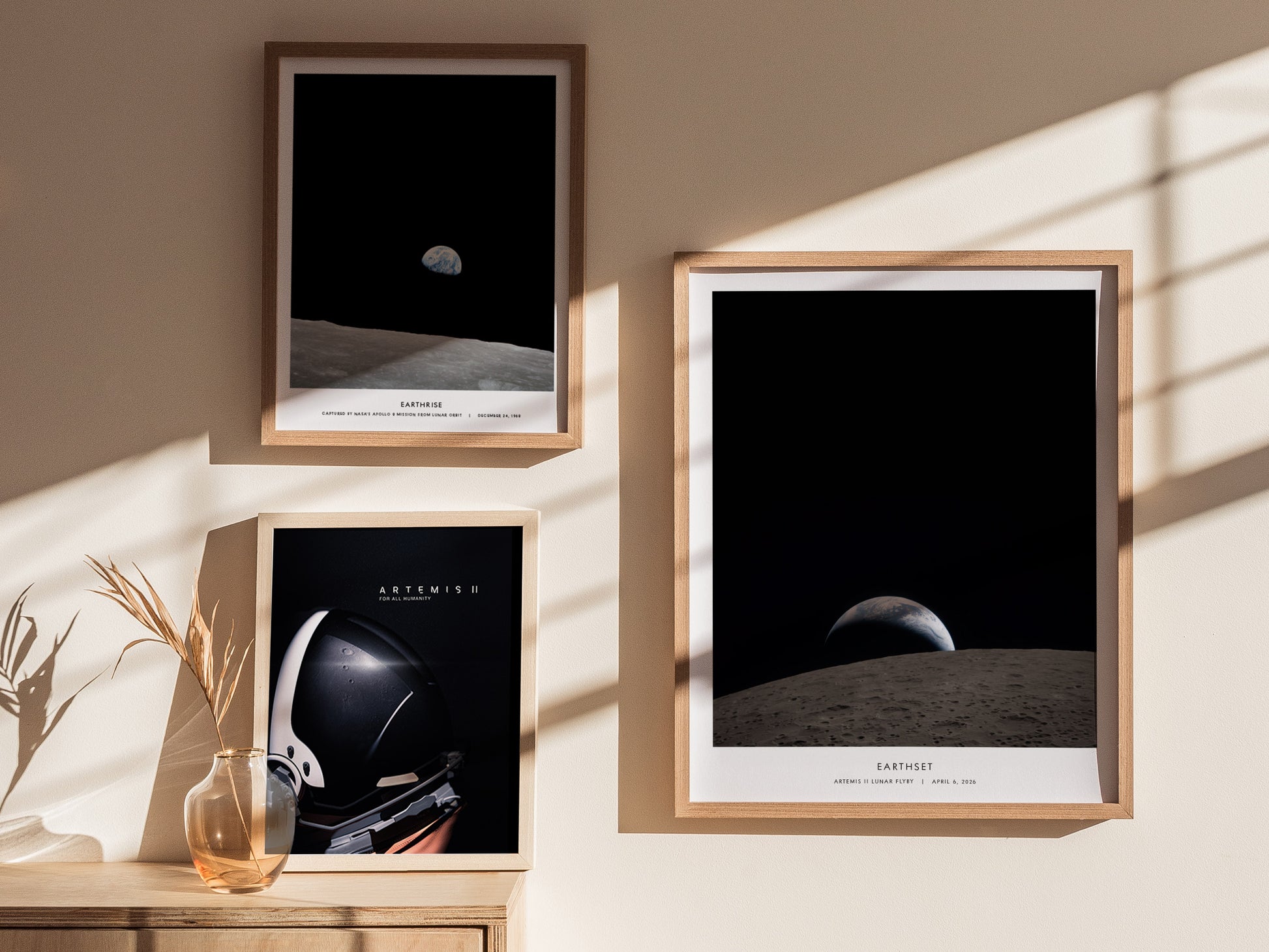 A stylish interior display featuring three framed posters on a light wall. The top two posters showcase celestial images of Earth from the Moon, while the lower poster features an astronaut helmet, all beautifully lit by natural sunlight.