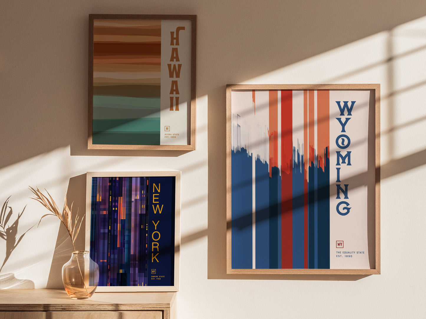 Three framed posters on a sunlit wall feature minimalist designs for Hawaii, Wyoming, and New York. Shadows from window panes add a dynamic touch to the scene.