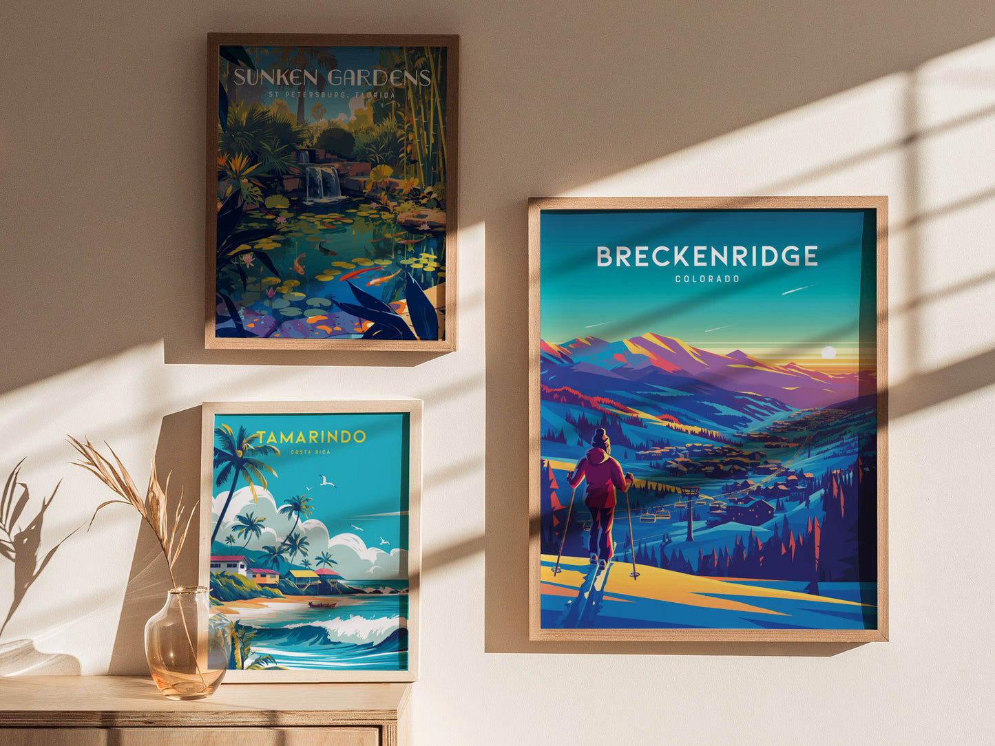 Three framed posters are displayed on a sunlit wall: Sunken Gardens, Tamarindo Costa Rica, and Breckenridge Colorado, featuring vibrant landscapes and scenes. A vase with dried plants sits below.