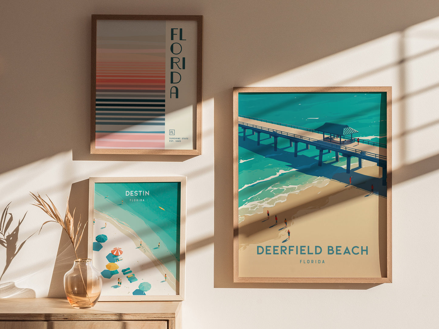Three framed posters are displayed on a sunlit wall: one of Deerfield Beach Pier, another of Destin, and a colorful striped Florida design. A vase with dried plants sits nearby.