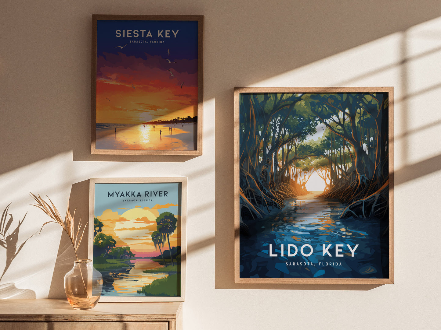 Three framed posters on a wall feature scenic illustrations of Siesta Key, Myakka River, and Lido Key in Sarasota, Florida. Sunlight casts shadows, enhancing the displays ambiance.