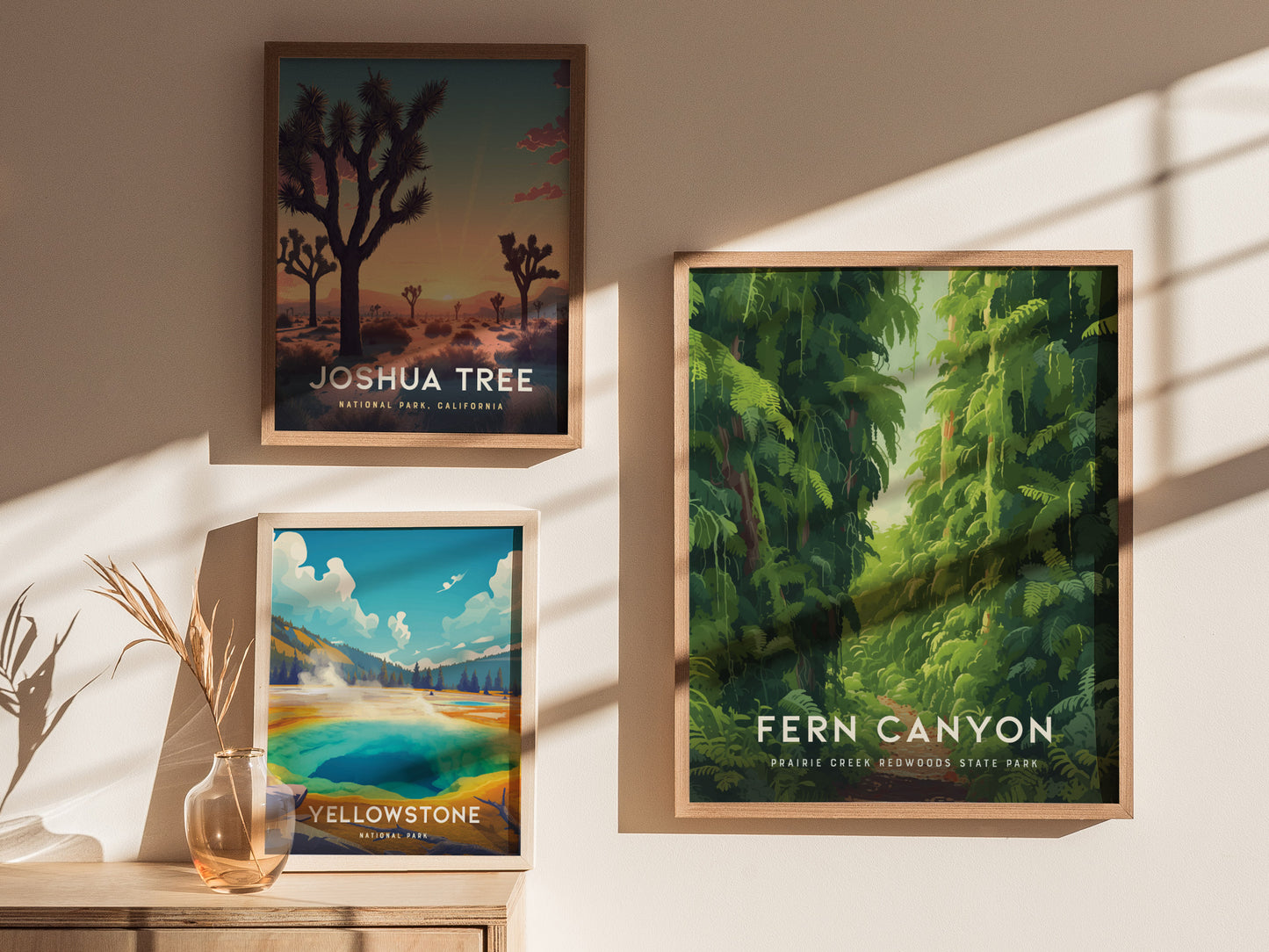 Three framed posters on a sunlit wall depict landscapes: Joshua Tree at sunset, Yellowstones vibrant geysers, and Fern Canyons lush greenery. A vase with dried plants sits below.