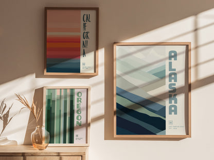 Three framed minimalist posters of California, Oregon, and Alaska are displayed on a beige wall with sunlight casting shadows. A small vase with dried grass sits on a wooden surface.