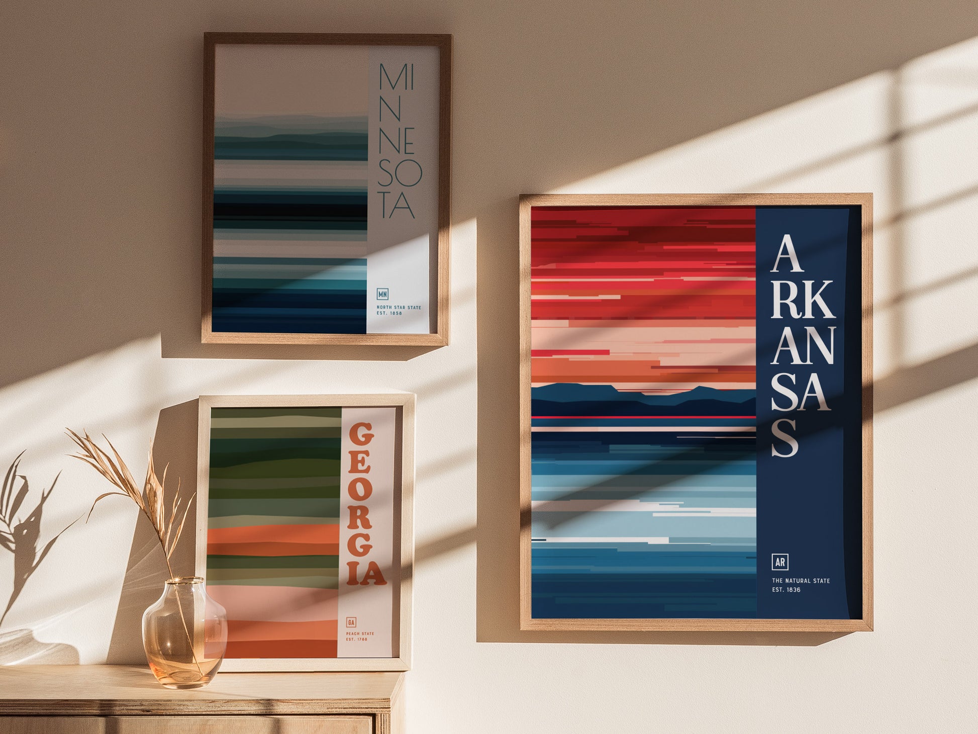 Three minimalist design posters for Minnesota, Georgia, and Arkansas are displayed on a wall. Each features abstract horizontal stripes in various colors, framed with wooden borders.