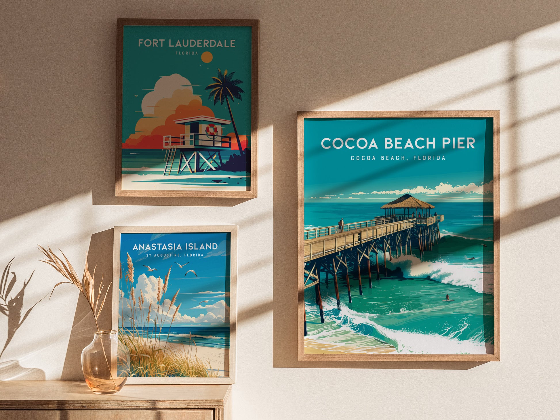 Three framed posters on a sunlit wall depict beach scenes from Fort Lauderdale, Anastasia Island, and Cocoa Beach Pier. A glass vase with dried plants sits on a wooden surface.