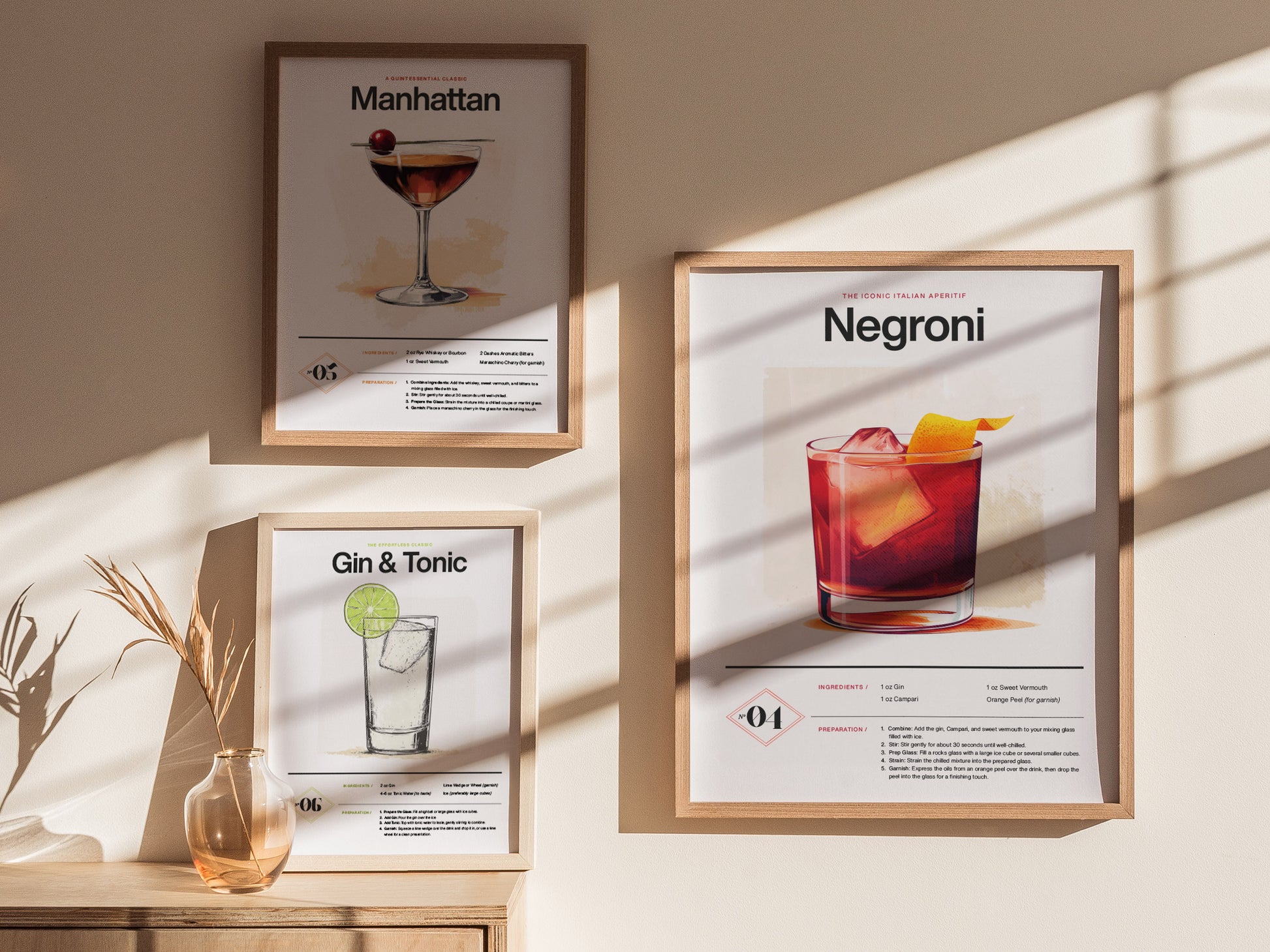 Three framed cocktail posters, Manhattan, Negroni, and Gin & Tonic, are displayed on a light-colored wall. Sunlight casts shadows, and a vase with dried plants sits below.