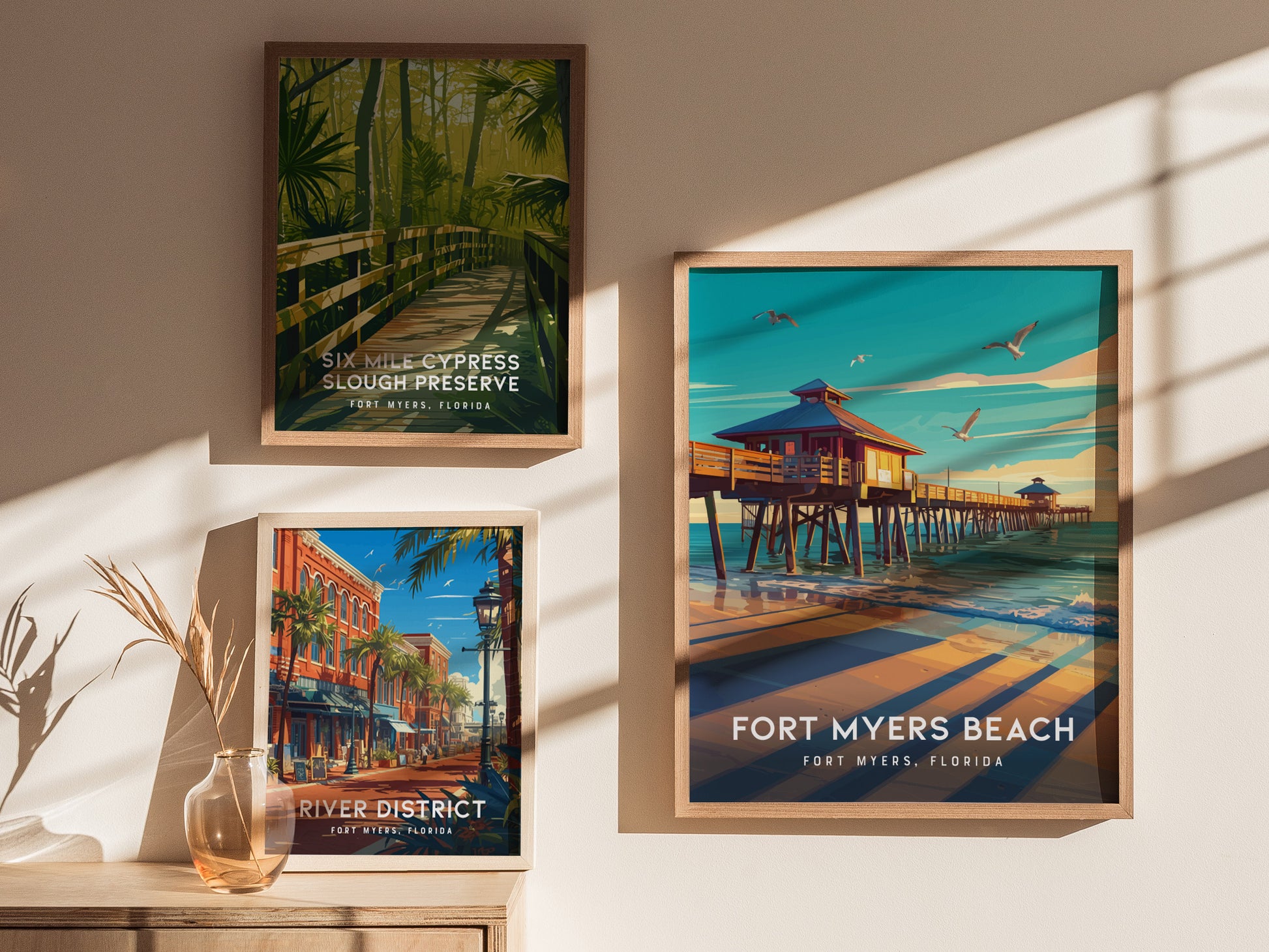 Three framed posters are displayed on a wall, featuring Fort Myers Beach pier, a lush boardwalk in Six Mile Cypress Slough Preserve, and a vibrant street in the River District.