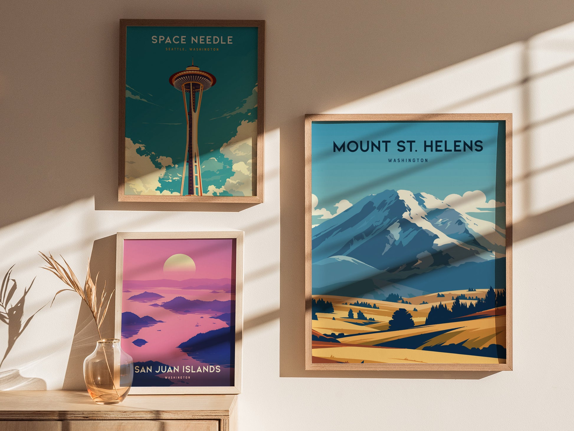 Three framed posters are displayed on a wall, featuring the Space Needle, Mount St. Helens, and San Juan Islands, with soft sunlight casting shadows across them.