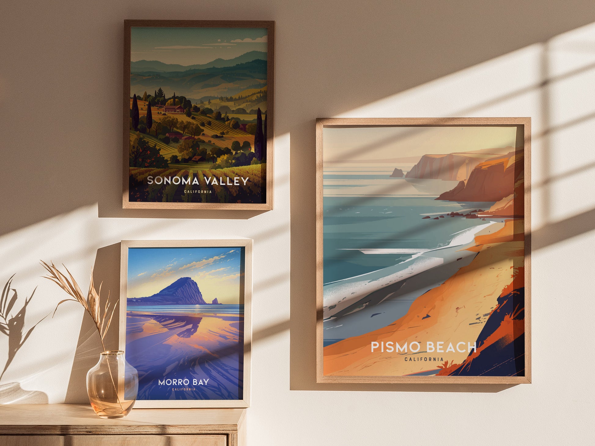 Framed posters of Sonoma Valley, Morro Bay, and Pismo Beach are displayed on a sunlit wall. A glass vase with dried grass accents the wooden surface below the posters.