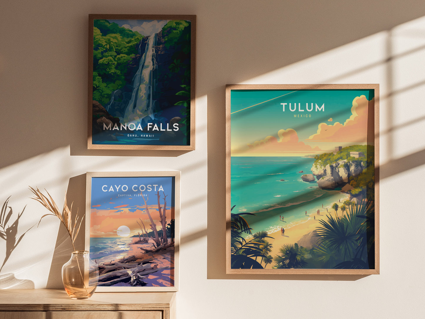Three framed posters are displayed on a wall, featuring Manoa Falls, Tulum, and Cayo Costa landscapes. A glass vase with dried plants sits on a wooden surface below.