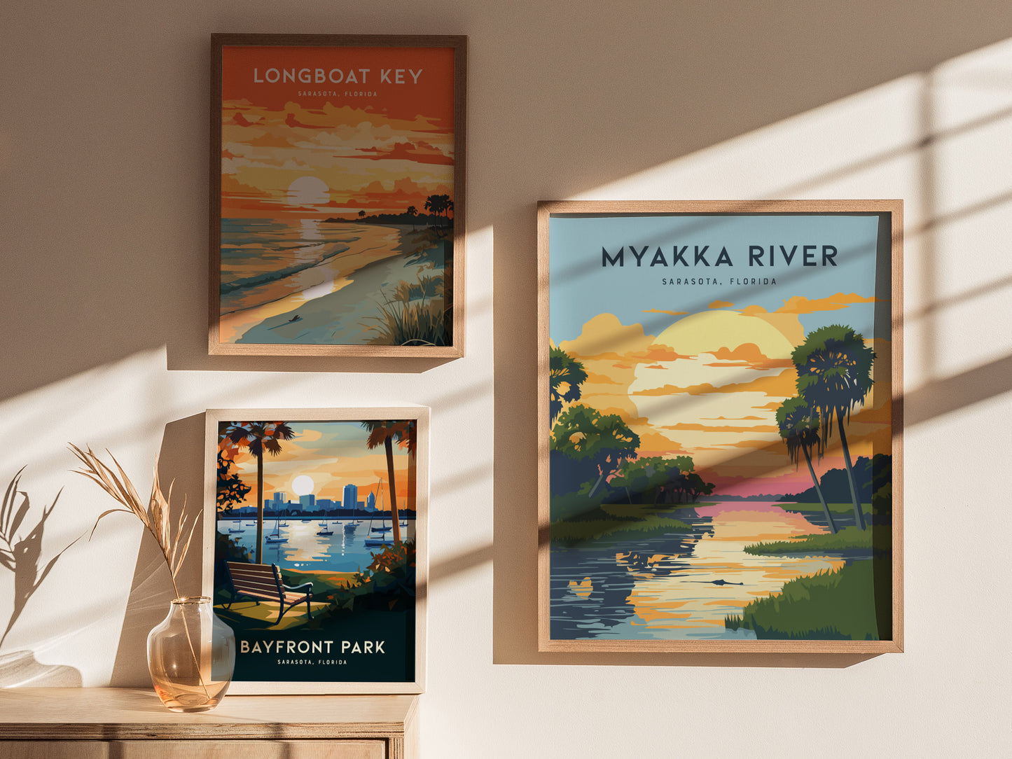 Three framed posters on a beige wall depict scenic views of Sarasota, Florida: Myakka River, Longboat Key, and Bayfront Park, with sunlight casting shadows nearby.
