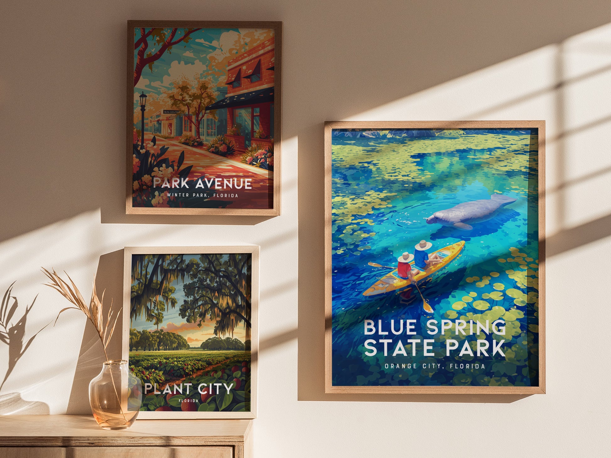 Three framed posters are displayed on a wall with sunlight casting shadows. The featured poster highlights Blue Spring State Park with kayakers and a manatee in clear waters.