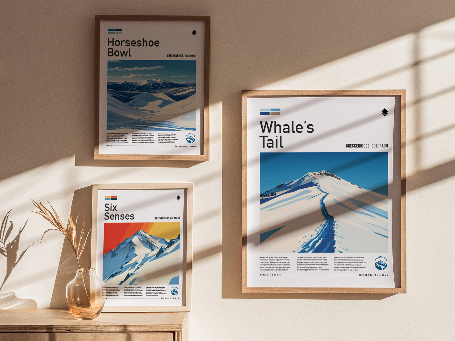 Three framed posters on a wall depict ski runs, titled Whales Tail, Horseshoe Bowl, and Six Senses, with scenic snowy mountain landscapes. A vase with dried plants sits below.