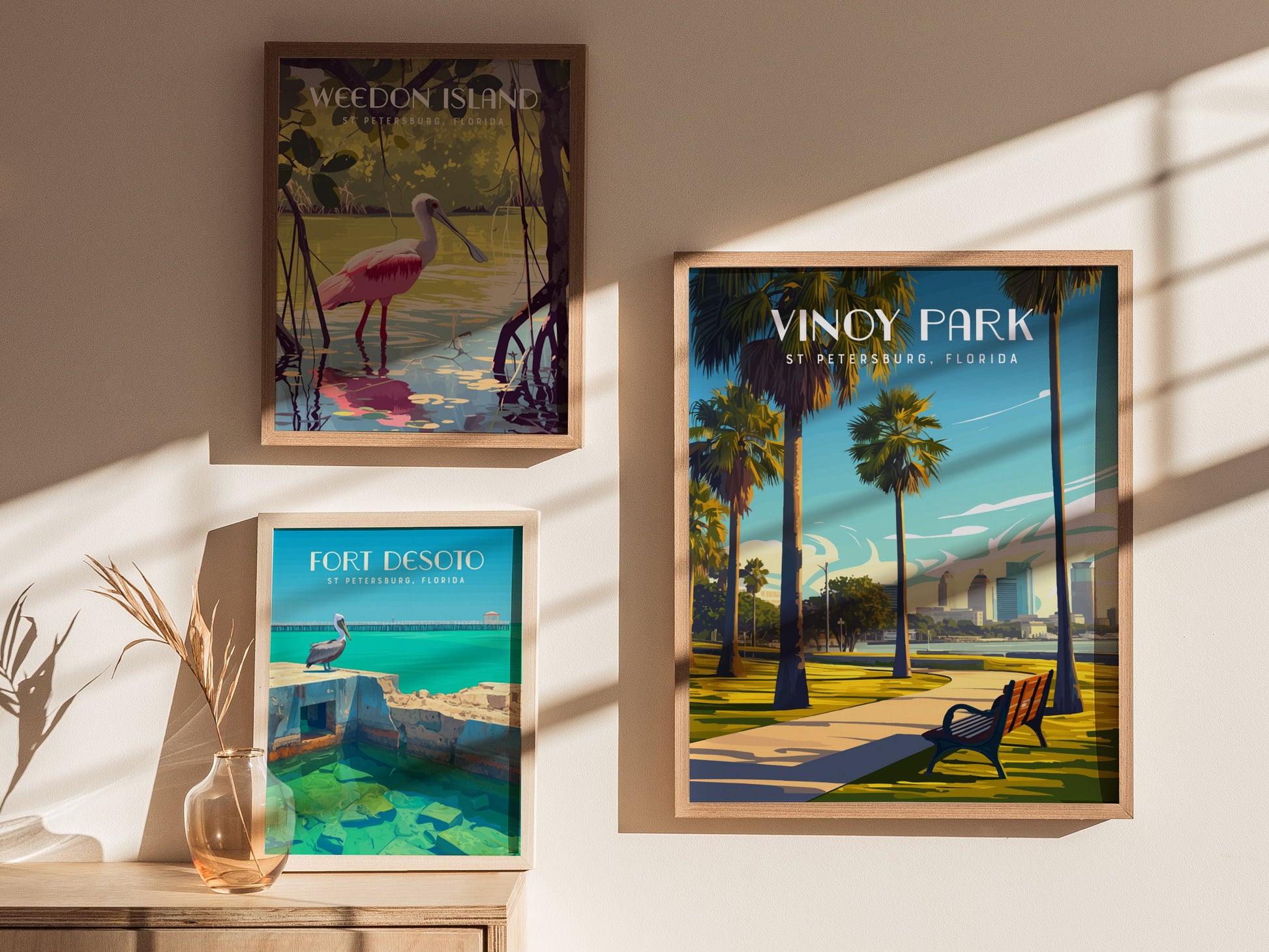 Framed posters of Weedon Island, Fort Desoto, and Vinoy Park are displayed on a sunlit wall. A vase with dried grass sits on a wooden surface below.