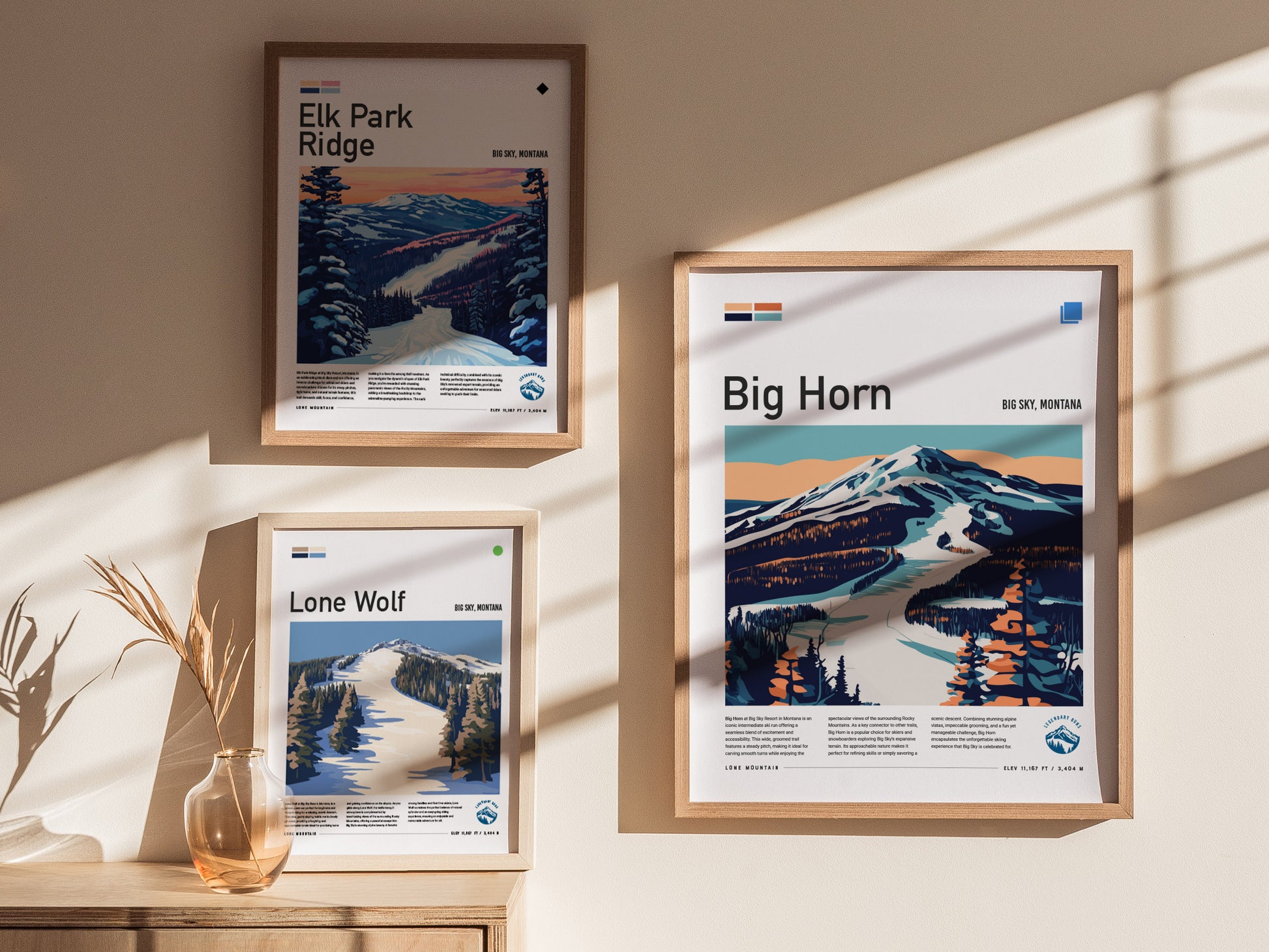 Three framed posters featuring stylized illustrations of ski runs in Big Sky, Montana, are displayed on a sunlit wall. A small vase with dried plants sits on a table.