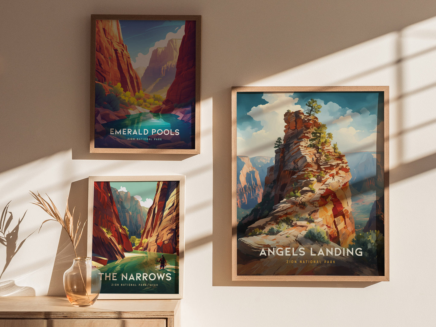 Three framed posters depicting Zion National Park landscapes, including Angels Landing, Emerald Pools, and The Narrows, are displayed on a sunlit wall above a wooden surface with a vase.