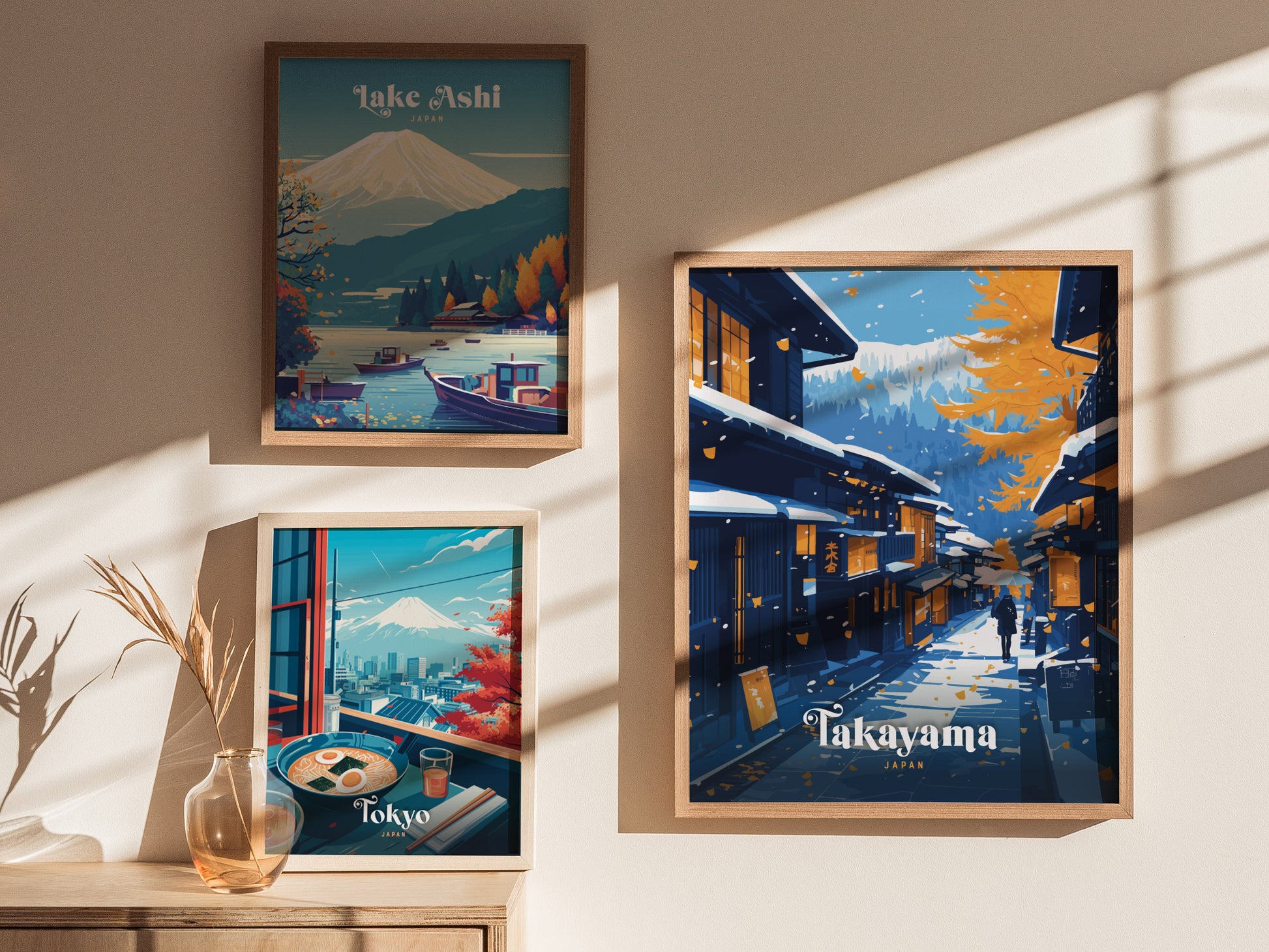 Framed posters on a wall depict Lake Ashi, Tokyo with Mount Fuji, and a snowy Takayama street scene. Sunlight casts shadows across the wall and a vase with dried grass.
