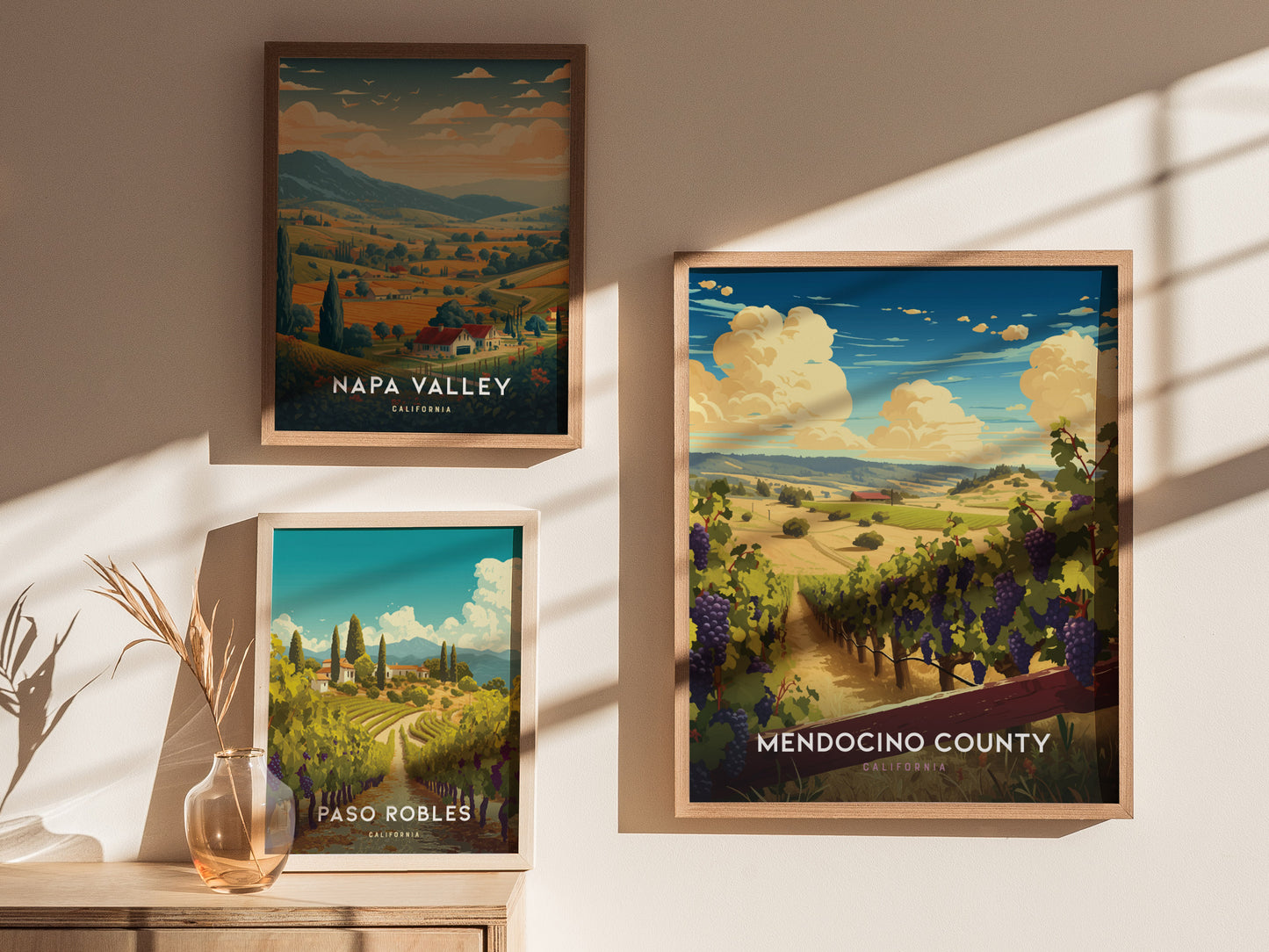 Framed posters of Napa Valley, Paso Robles, and Mendocino County are displayed on a sunlit wall, featuring vibrant vineyard and countryside landscapes. A vase with dried grass sits below.