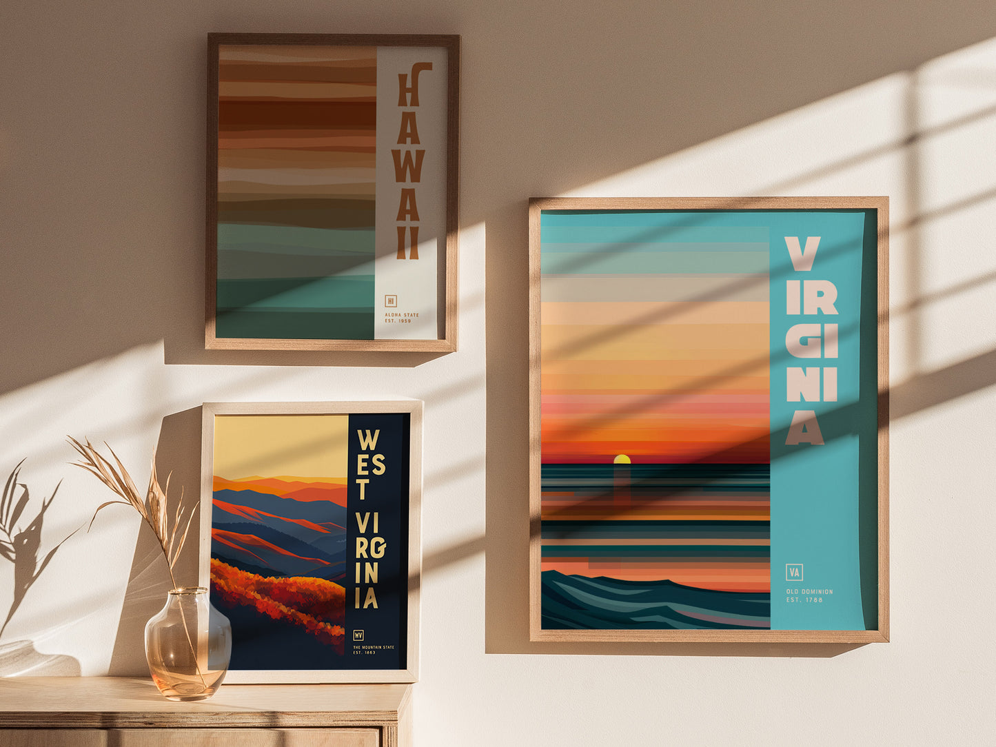 Three framed posters on a wall display minimalist designs representing Hawaii, Virginia, and West Virginia. Sunlight casts shadows over the vibrant colors and clean typography.