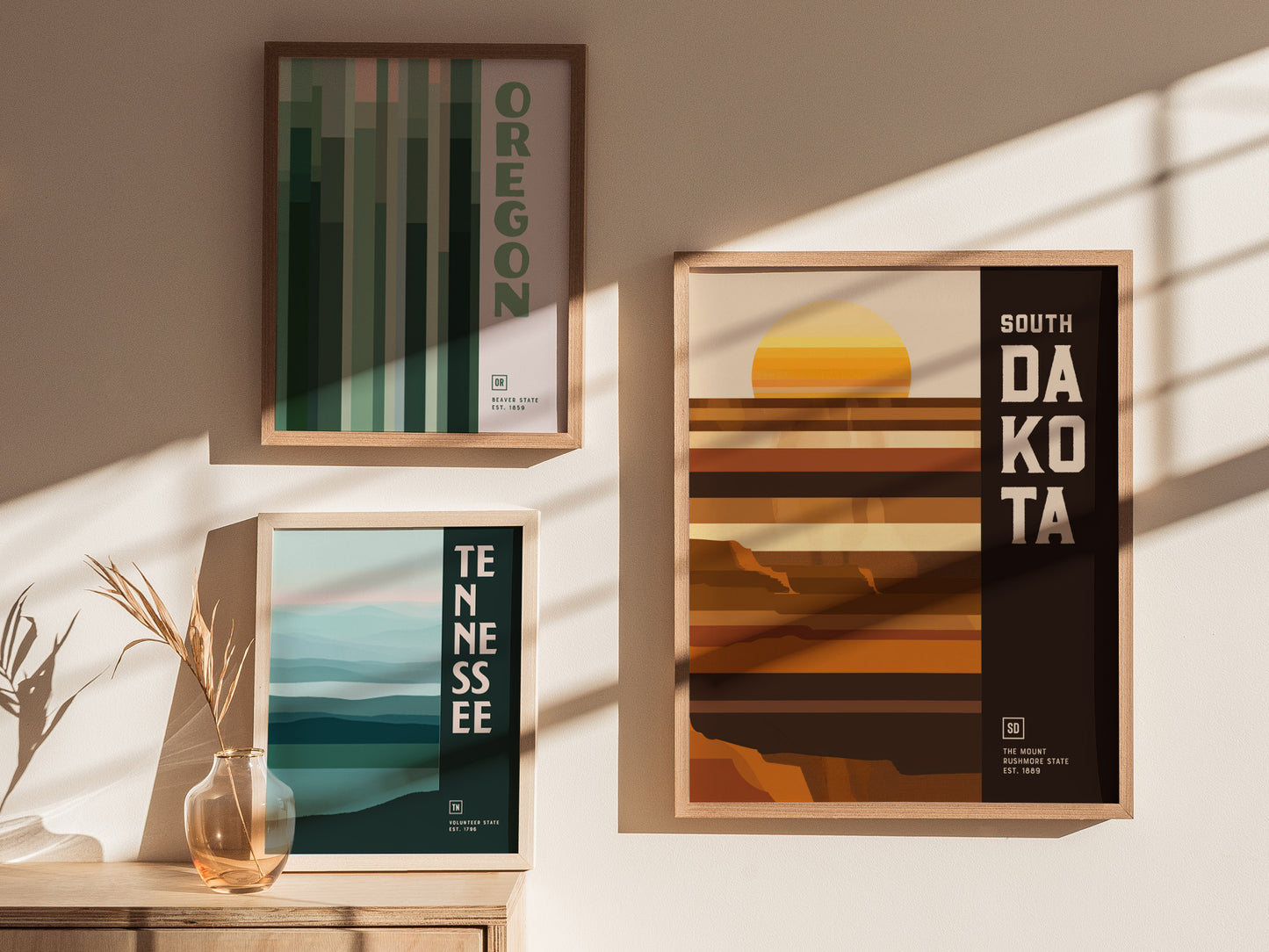 Three framed posters are displayed on a wall, featuring geometric designs for Oregon, Tennessee, and South Dakota. Sunlight casts soft shadows across the minimalist, earthy-toned artwork.