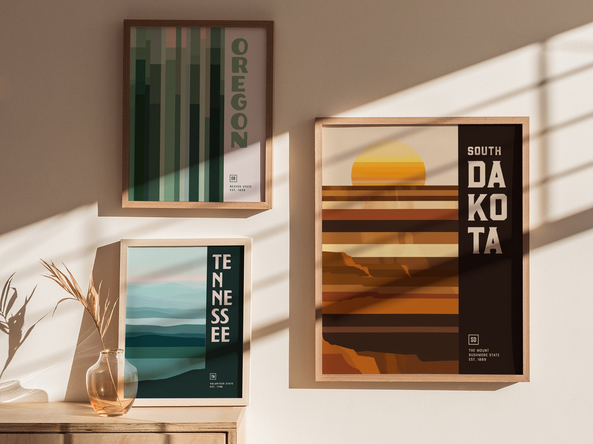 Three framed posters are displayed on a wall, featuring geometric designs for Oregon, Tennessee, and South Dakota. Sunlight casts soft shadows across the minimalist, earthy-toned artwork.