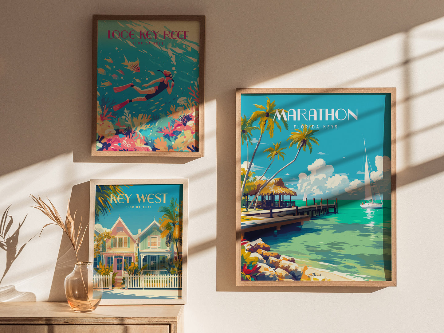 Three framed posters on a wall depict Florida Keys scenes: a diver near coral, colorful beach houses, and a tropical pier with palm trees. Sunlight casts shadows across them.