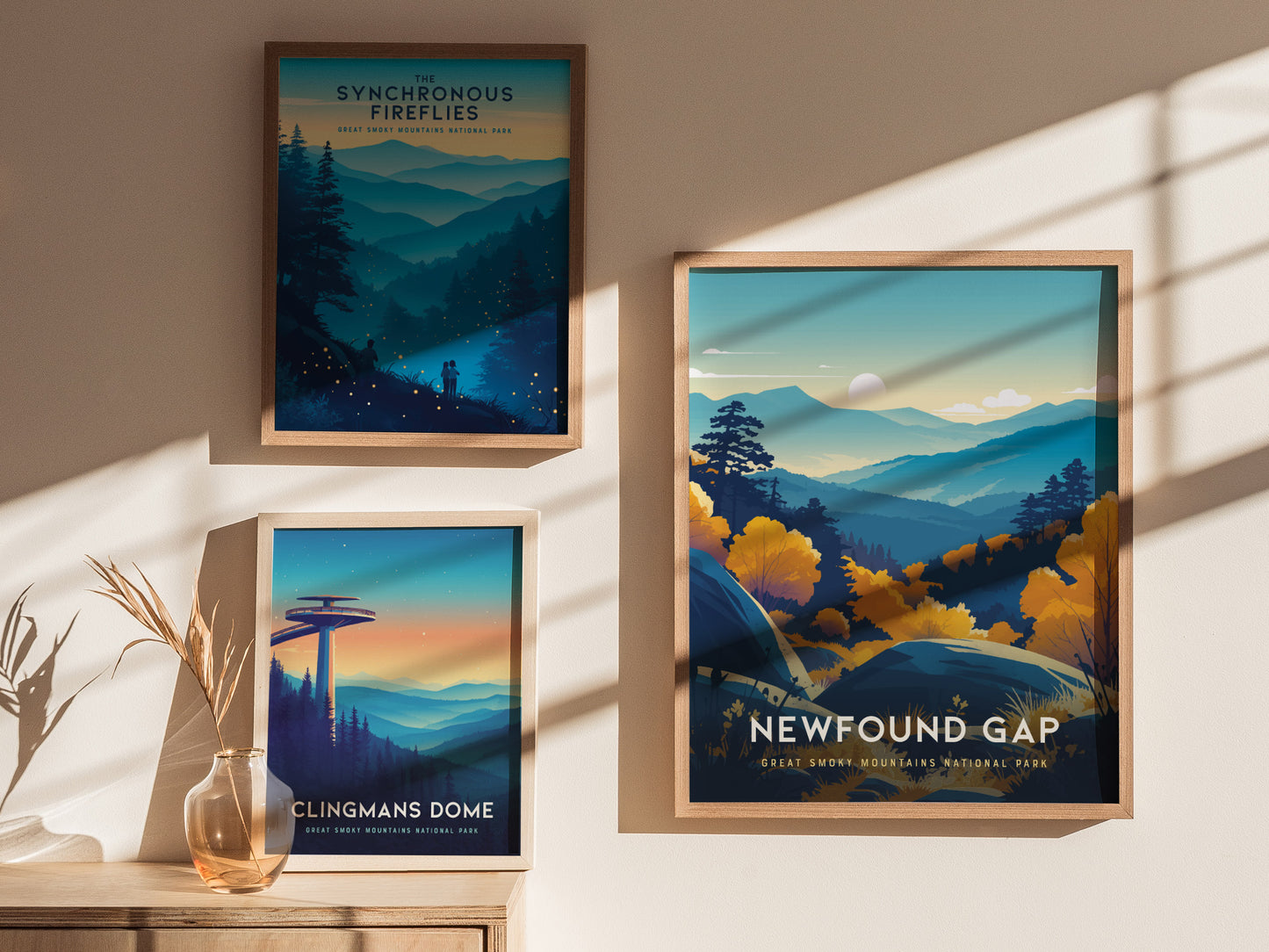 Three framed posters hang on a wall, each depicting scenic views of the Great Smoky Mountains. The Newfound Gap poster features vibrant blue and yellow tones with layered peaks and trees. Sunlight creates shadows on the wall.
