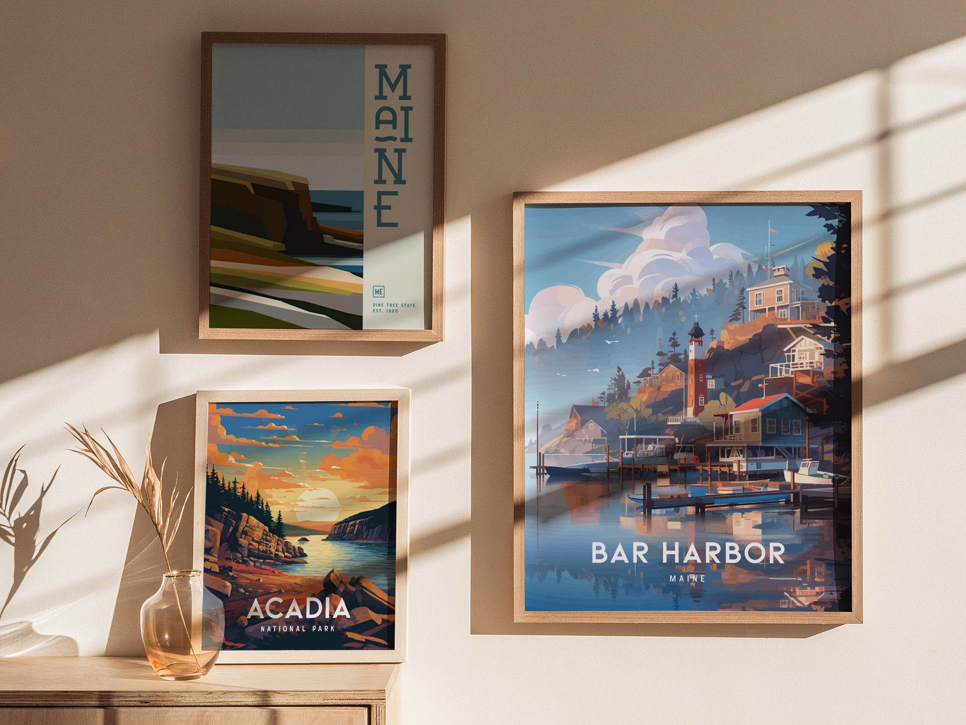 Three framed posters on a wall depict Maine landscapes, including Bar Harbor and Acadia National Park, with a vase holding dried plants on a wooden surface below.