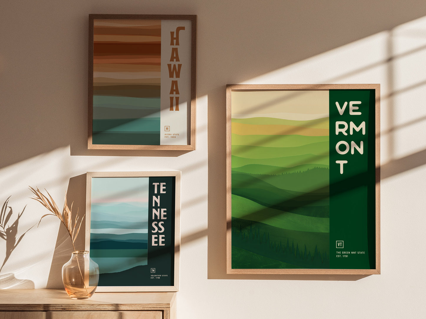 Three framed posters of Hawaii, Tennessee, and Vermont with abstract landscape designs hang on a wall. Sunlight casts shadows, and a vase with dried plants sits below.