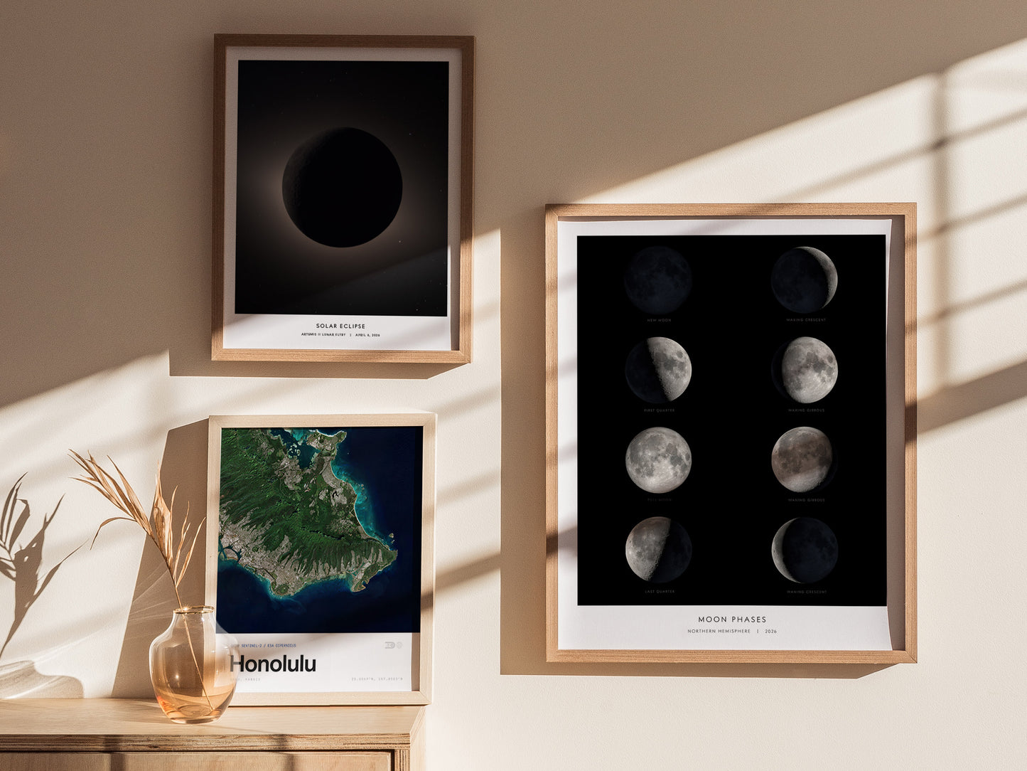A well-lit interior showcases three framed prints on a wall. The top left features a dark image of a solar eclipse, while the top right displays the eight lunar phases in vivid detail. The bottom left print shows a colorful map of Honolulu.