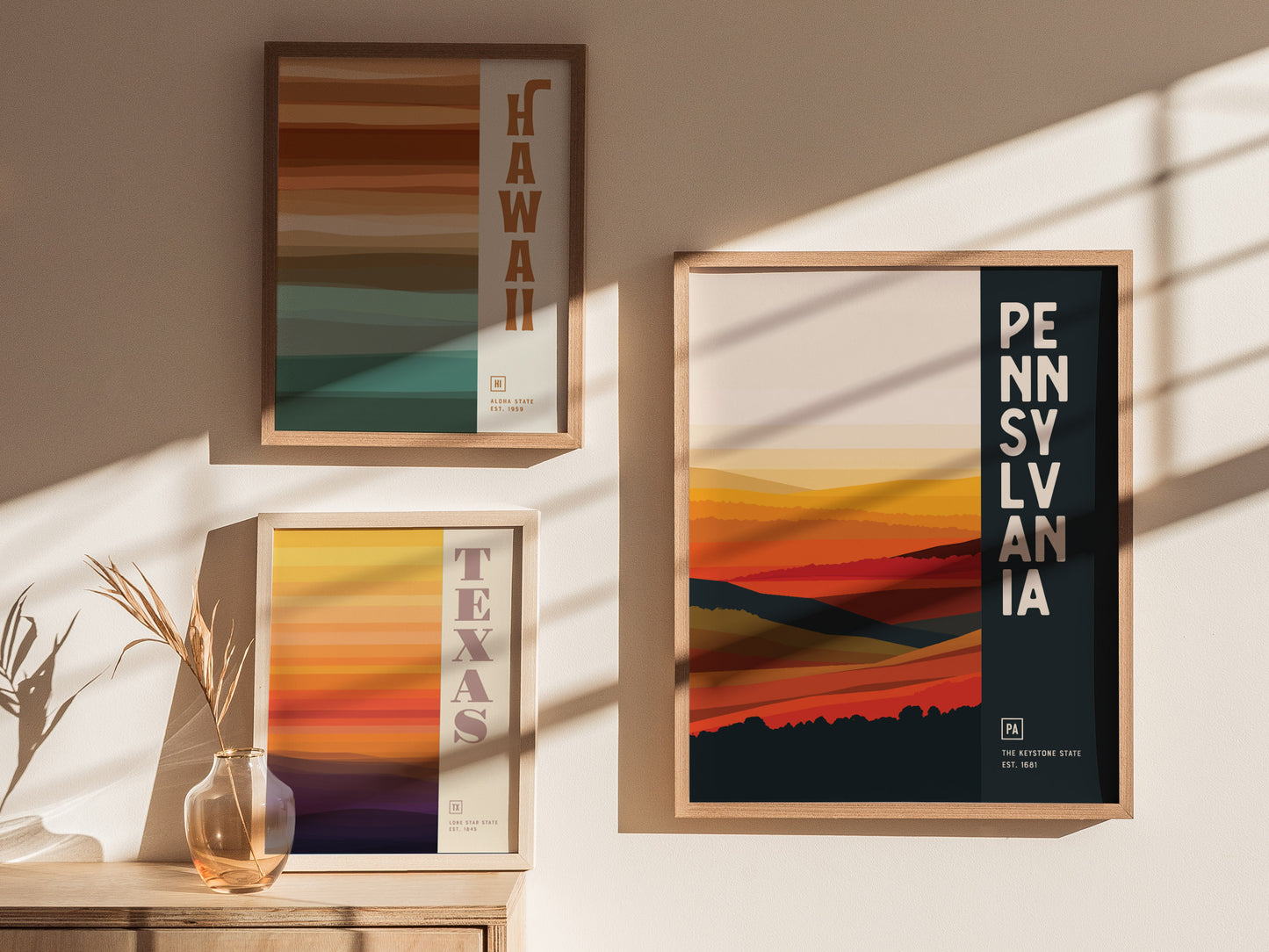 Three framed posters with abstract designs are displayed on a wall. They represent Hawaii, Texas, and Pennsylvania, featuring colorful horizontal stripes and bold text naming each state.
