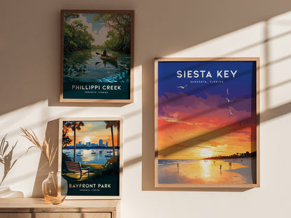 Three framed posters hang on a wall, featuring illustrations of Phillippi Creek, Bayfront Park, and a vibrant Siesta Key sunset. A vase with dried plants sits on a table.