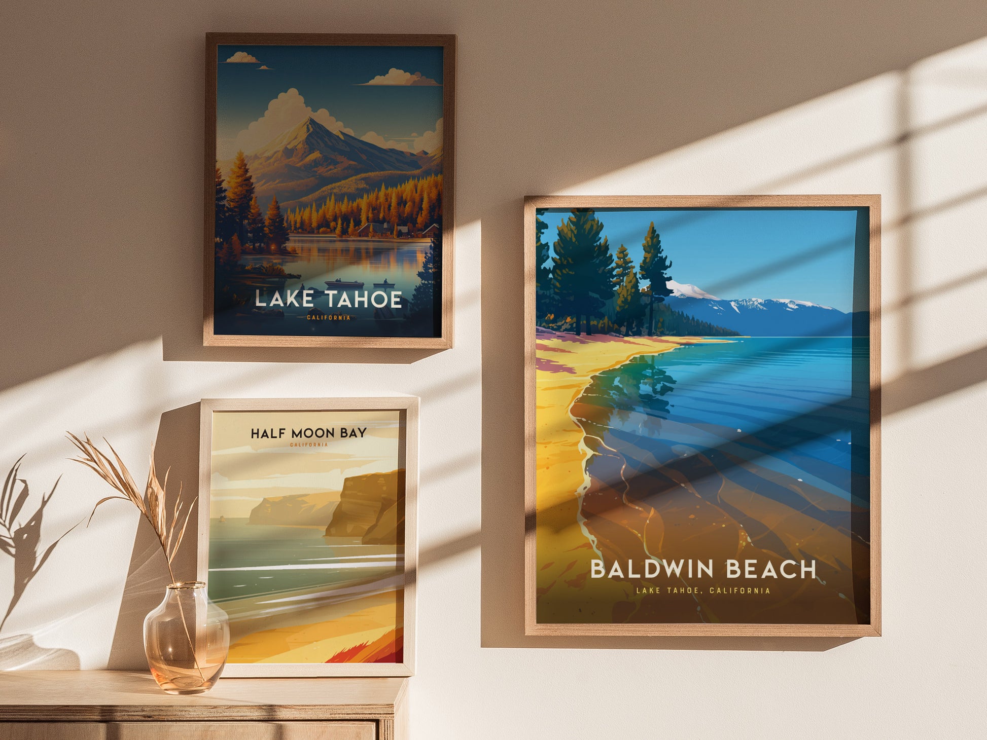 Three framed travel posters are displayed on a sunlit wall. They depict scenic landscapes of Lake Tahoe, Baldwin Beach, and Half Moon Bay, with a small vase nearby.