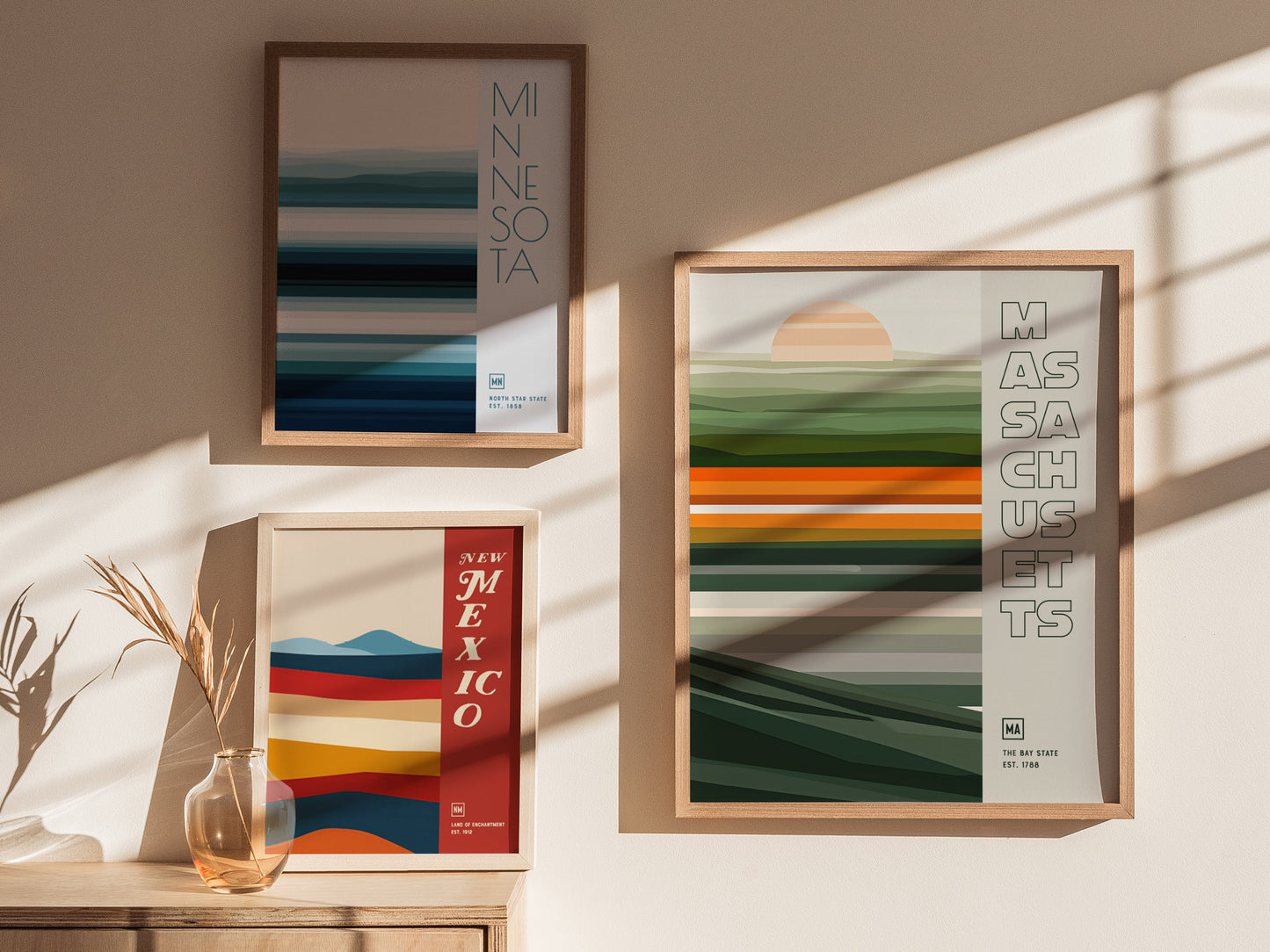 Three framed posters on a sunlit wall display minimalist designs for Minnesota, New Mexico, and Massachusetts, featuring horizontal color bands and text, with a vase of dried plants nearby.
