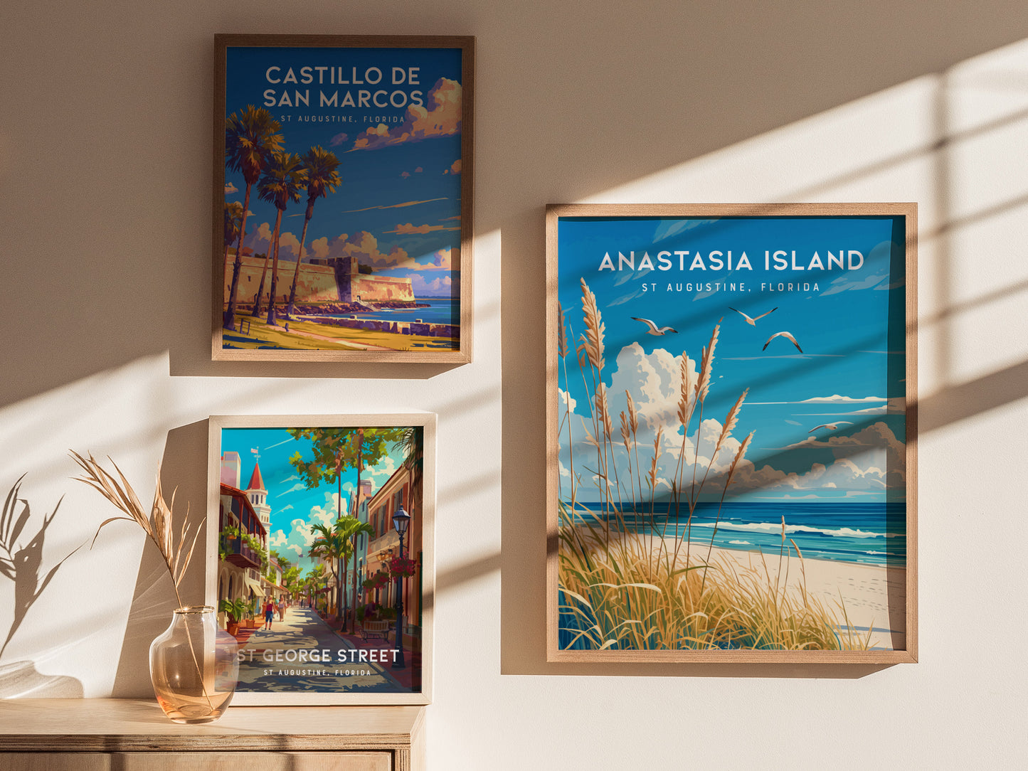 Three framed posters on a sunlit wall depict scenes from St. Augustine, Florida, including Castillo de San Marcos, Anastasia Island, and St. George Street, with a vase on a table.