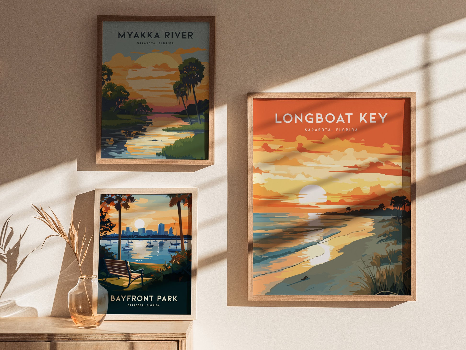 Three framed posters are displayed on a beige wall. The posters depict scenic views of Myakka River, Bayfront Park, and Longboat Key in Sarasota, Florida, featuring vibrant sunsets and natural landscapes.