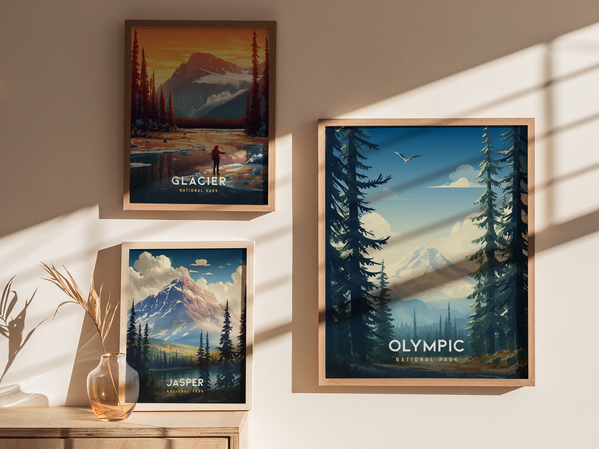Three framed posters are displayed on a wall, featuring scenic illustrations of Glacier, Jasper, and Olympic National Parks, with natural light casting shadows. A vase with dried plants is nearby.