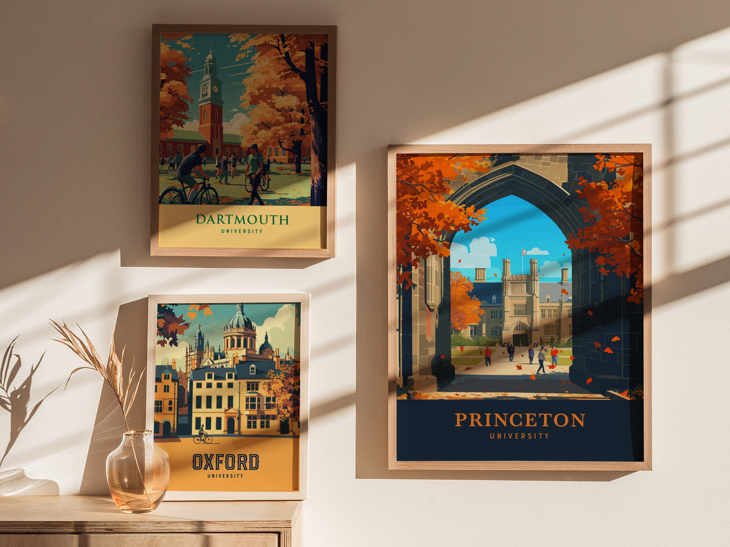 Three framed university posters are displayed on a sunlit wall, featuring Dartmouth, Oxford, and Princeton, each with vibrant autumn scenes and architectural landmarks. A vase with dried plants sits nearby.