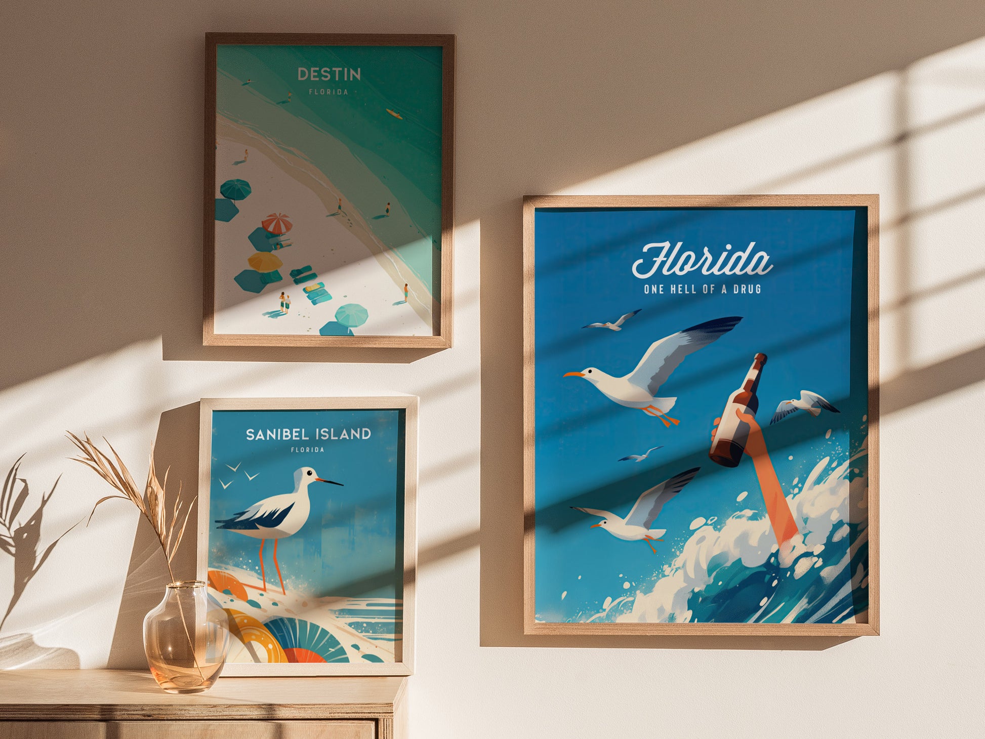 Three framed posters hang on a sunlit wall. The largest poster features seagulls, a bottle, and the text Florida: One Hell of a Drug against a blue background.