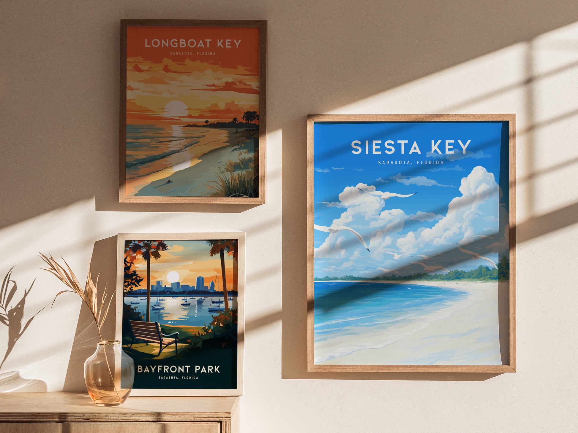 Three framed posters hang on a wall, showcasing vibrant beach scenes of Longboat Key, Siesta Key, and Bayfront Park. Sunlight casts soft shadows across the artwork.