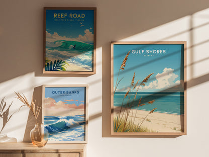 Three framed posters are displayed on a sunlit wall, depicting coastal scenes from Reef Road, Gulf Shores, and Outer Banks. A glass vase with dried grass sits nearby.